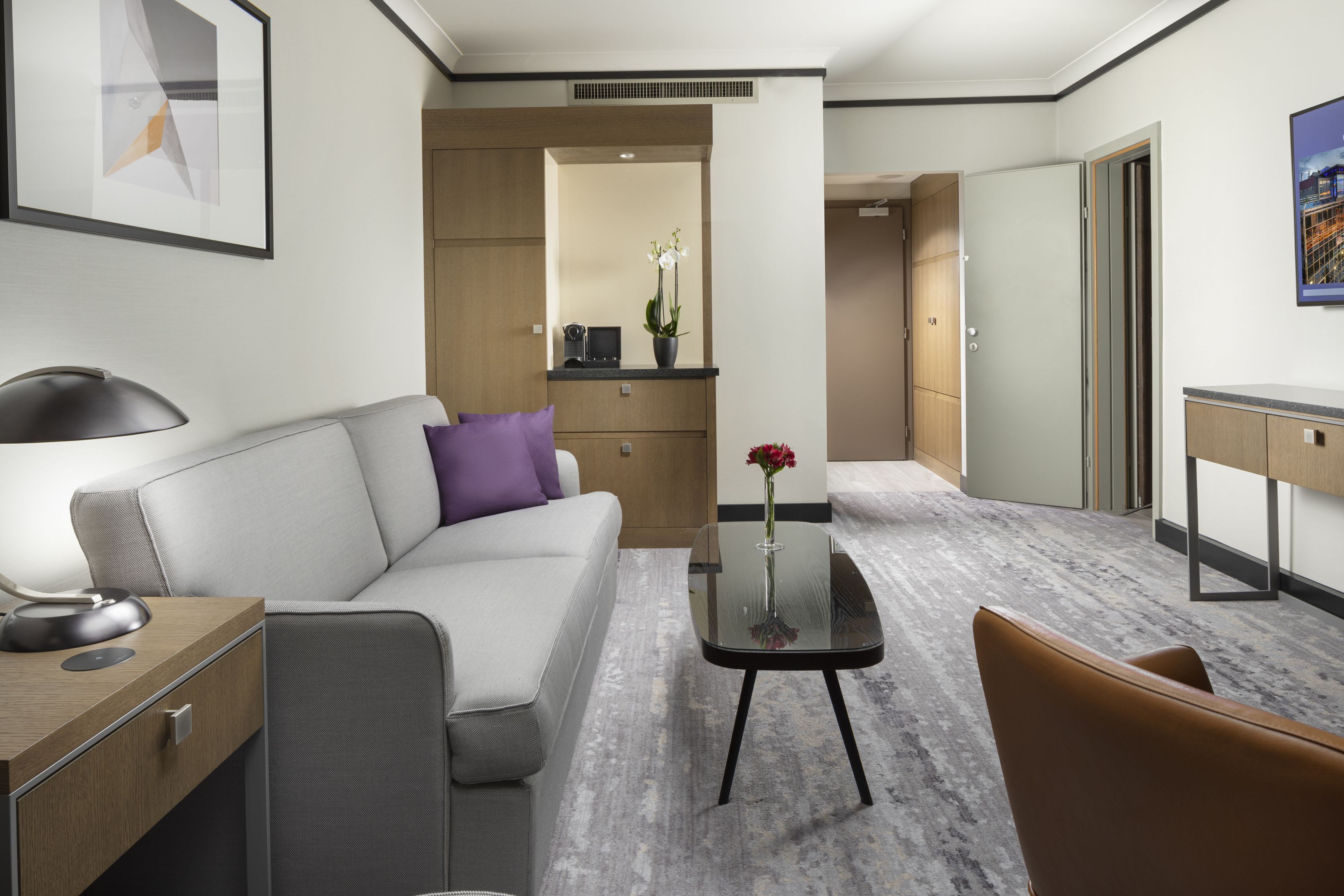 Et moderne hotelværelse med en grå sofa, et lille bord med en vase blomster, et skrivebord med lampe og et garderobeskab.