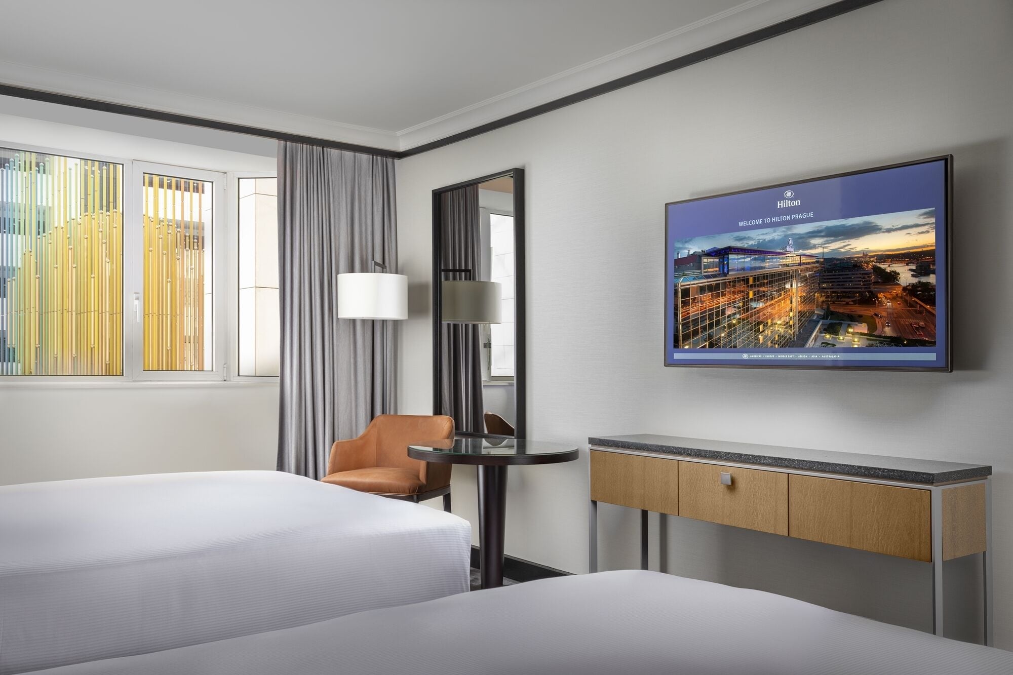A hotel room with two beds, a desk, a chair, a large window with curtains, and a cityscape poster on the wall.