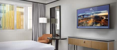 A hotel room with two beds, a desk, a chair, a large window with curtains, and a cityscape poster on the wall.