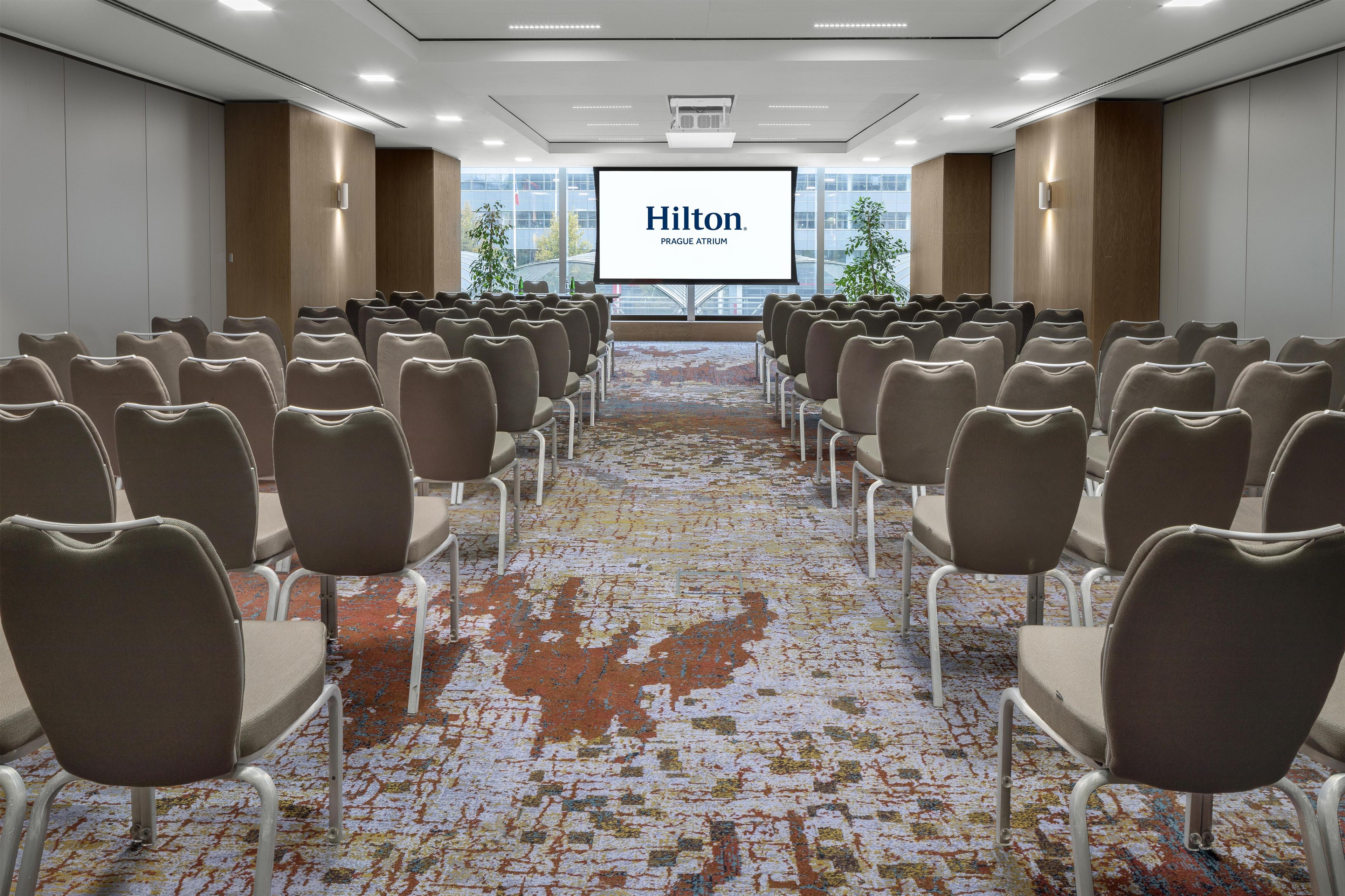 Un salón de conferencias con filas de sillas apuntando hacia una pantalla que muestra 'Hilton Prague Atrium'.