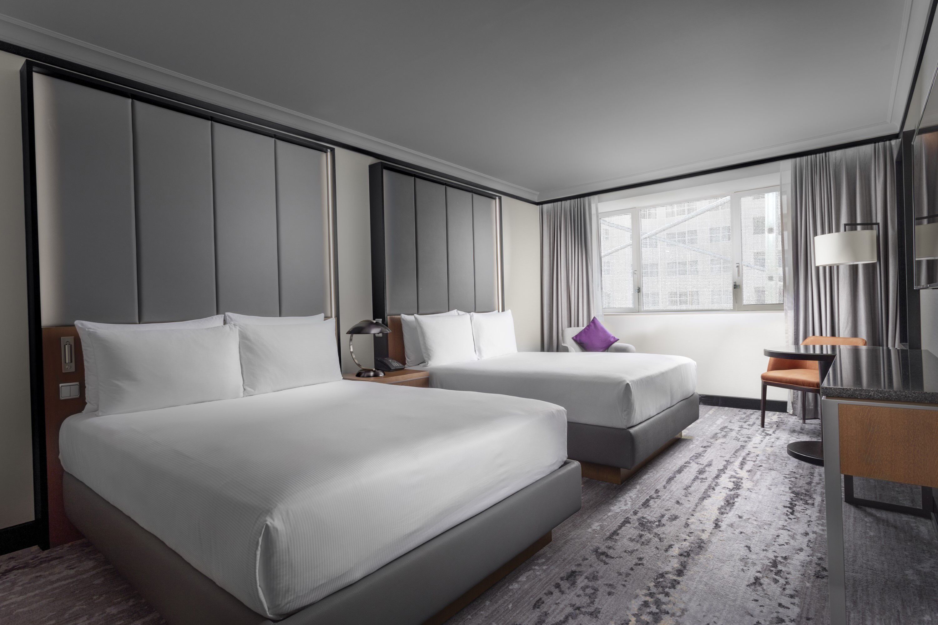 Habitación de hotel con dos camas, un escritorio, una silla, una lámpara y un ventanal con cortinas.