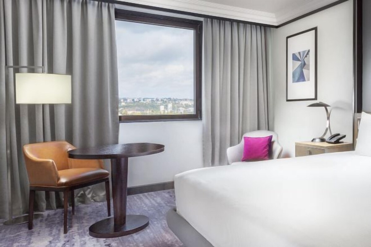 A hotel room with a bed, a chair, a small table, a lamp, and a view of the city through a window.