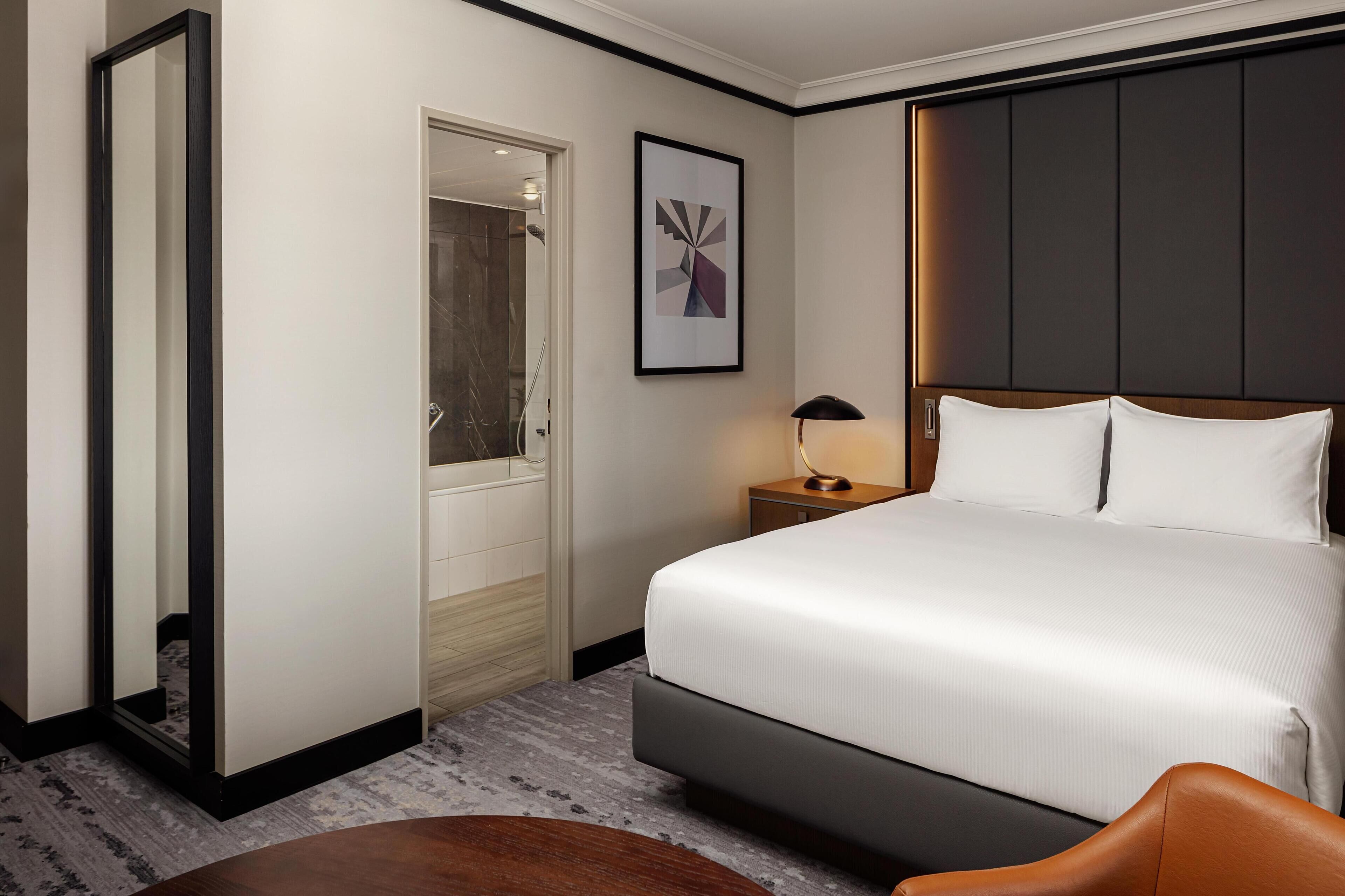 Una camera d'albergo con un letto grande, un comodino con una lampada, un quadro appeso al muro e una porta che conduce al bagno.
