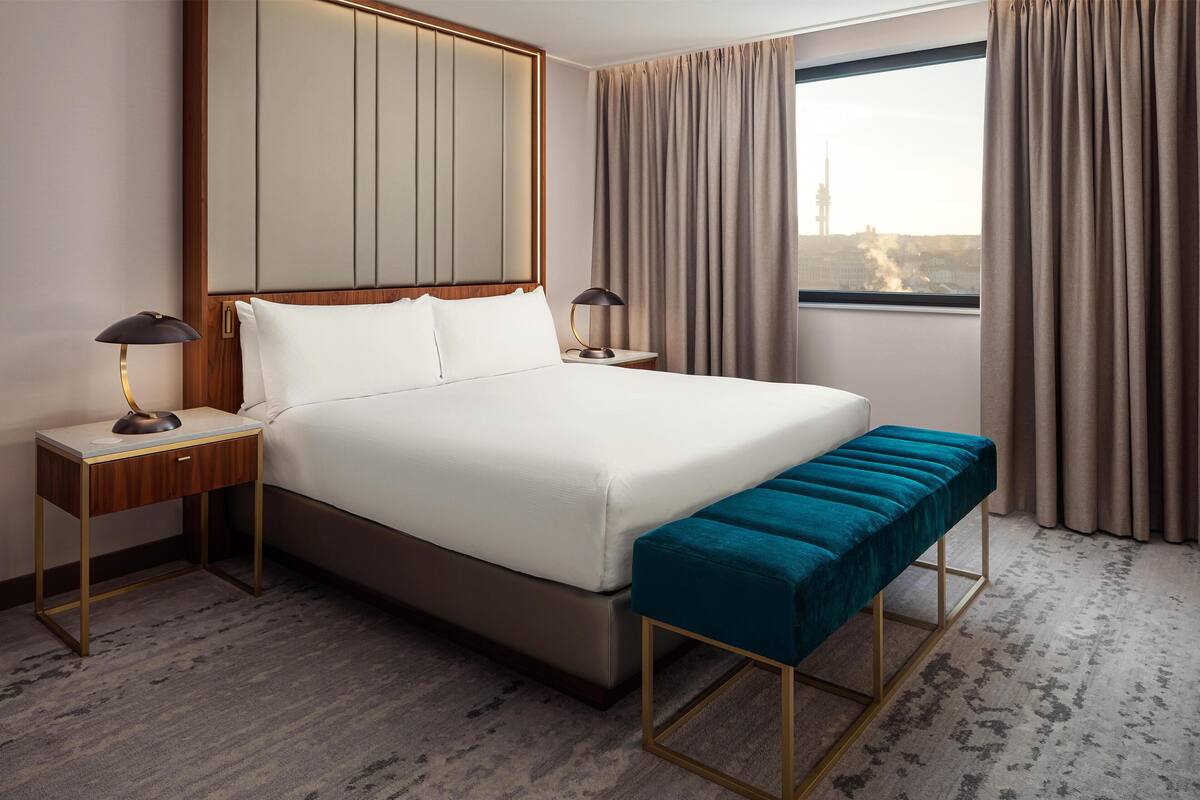 Ett hotellrum med en stor säng, två sängbord med lampor, en bänk och utsikt över stadsbilden genom fönstret.