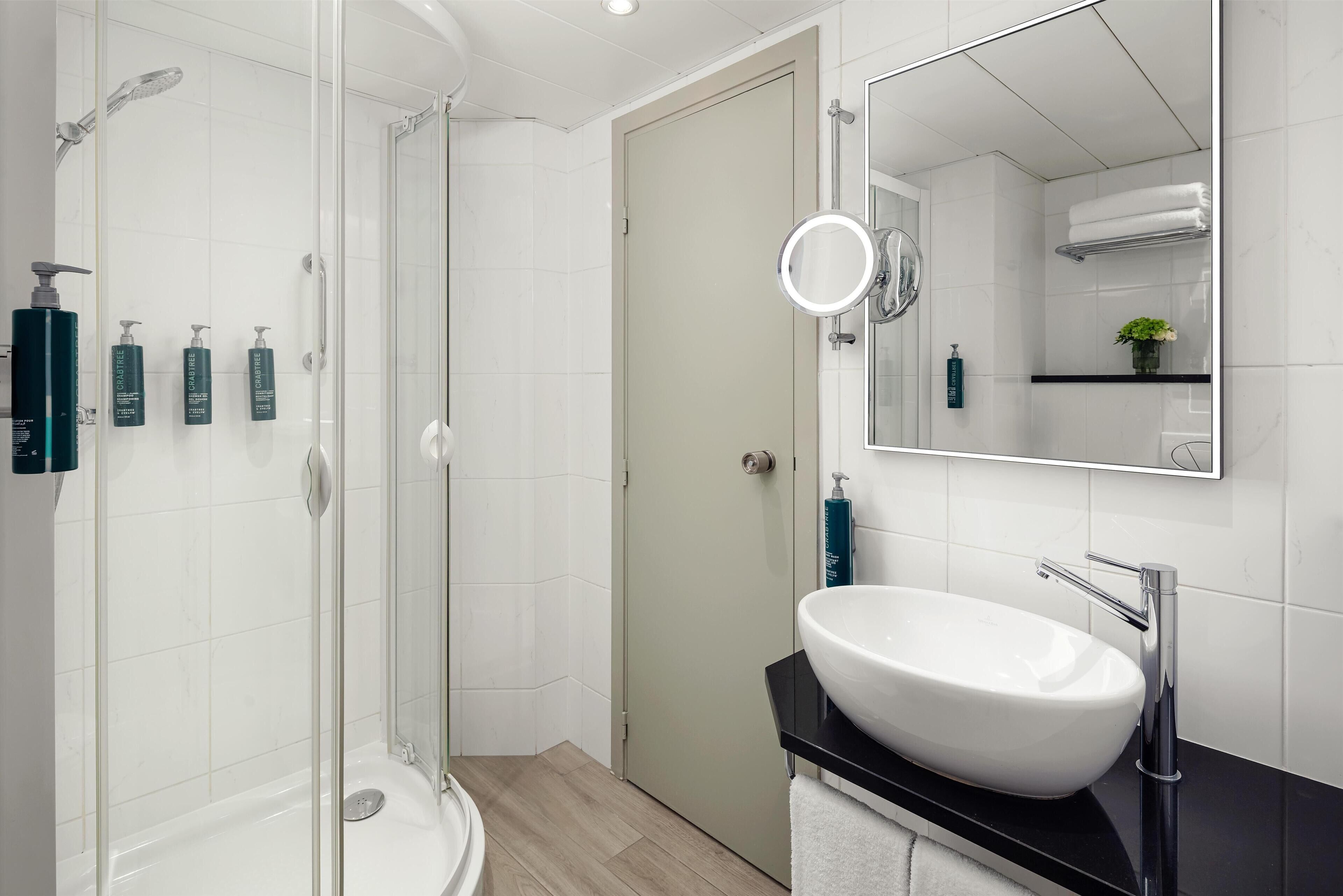 A modern bathroom with a glass-enclosed shower, a white sink with a black countertop, and a mirror with a round light.