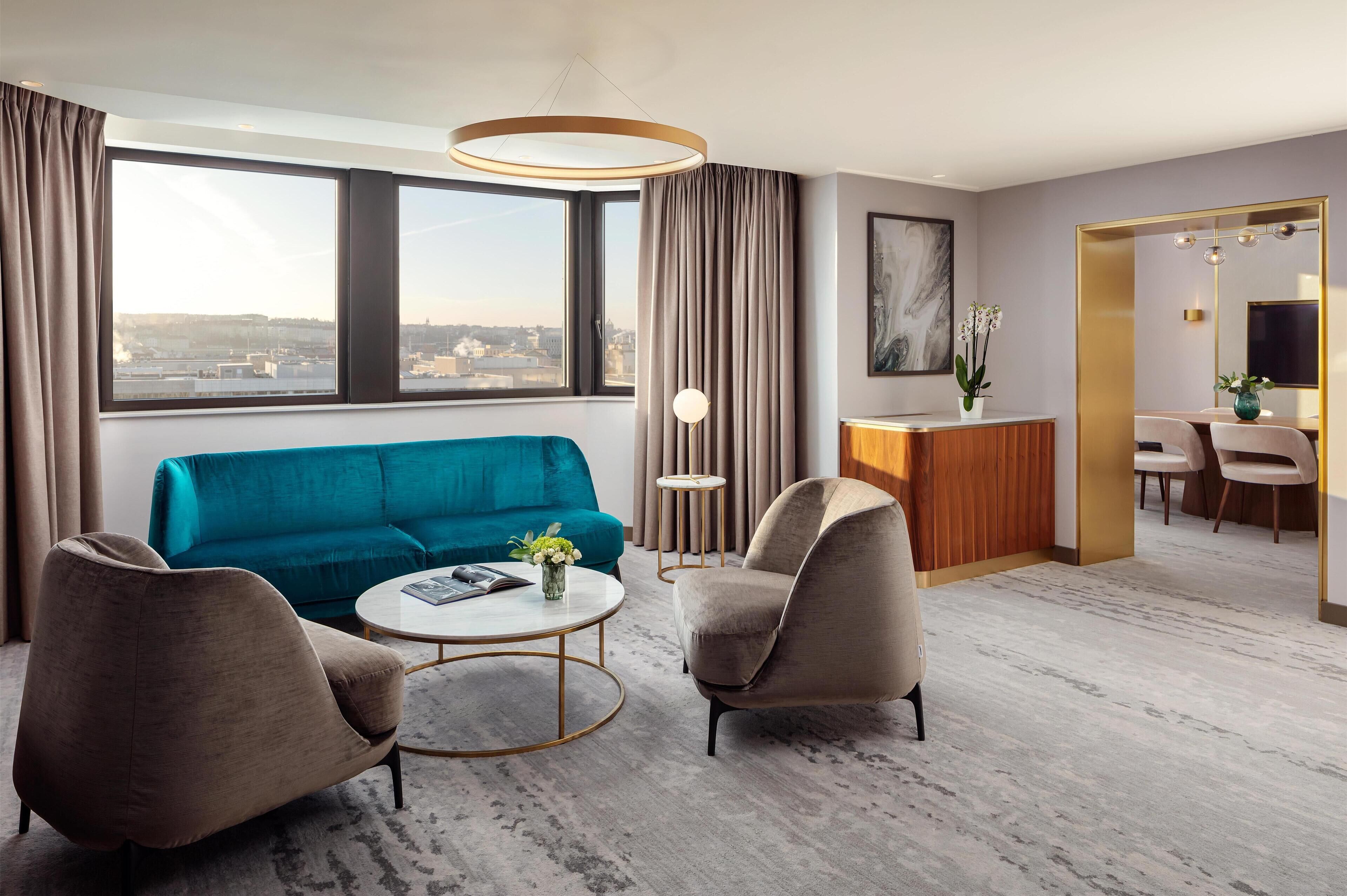 A modern hotel room with a teal sofa, two armchairs, a round coffee table, and a desk with a mirror.