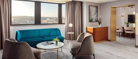 A modern hotel room with a teal sofa, two armchairs, a round coffee table, and a desk with a mirror.