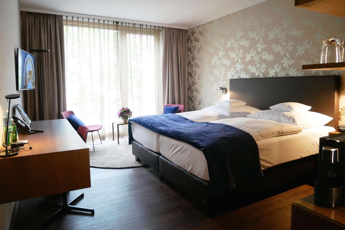 A hotel room with a bed, a desk with a telephone, a chair, a small table with a vase, and a TV on the wall.