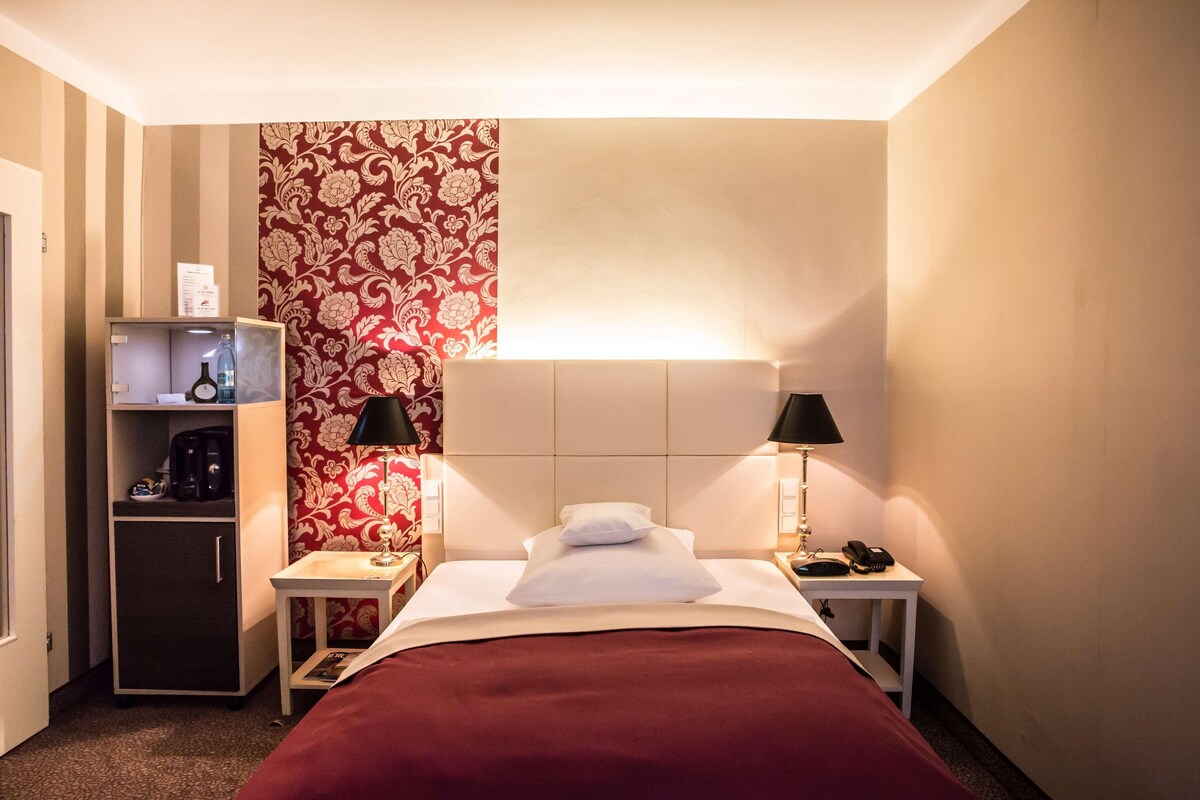 A hotel room with a bed, bedside tables, a nightstand, a wardrobe, and a red patterned wall.