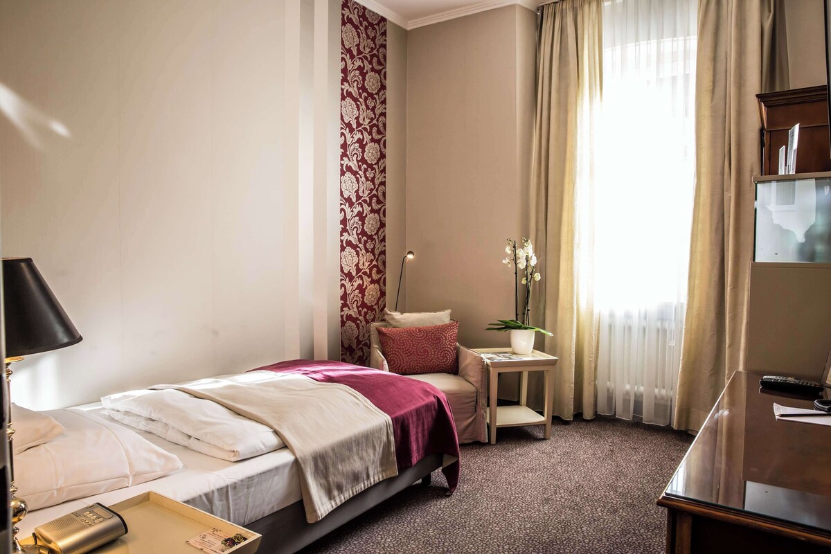 A hotel room with a bed, a desk, a chair, a small table with a plant, and a window with curtains.