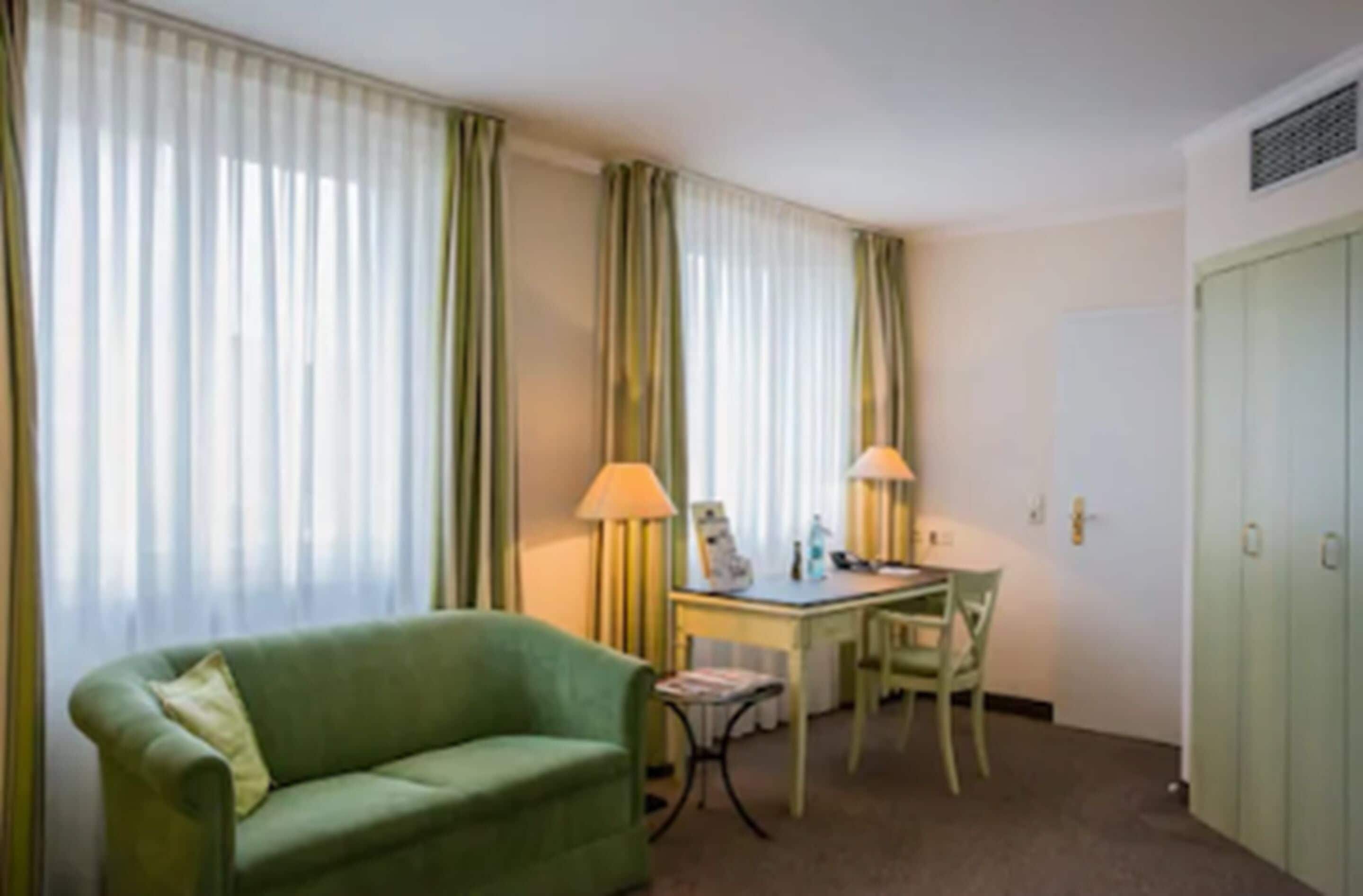 Quarto de hotel com um sofá verde, uma escrivaninha com abajur, uma cadeira e um guarda-roupa.