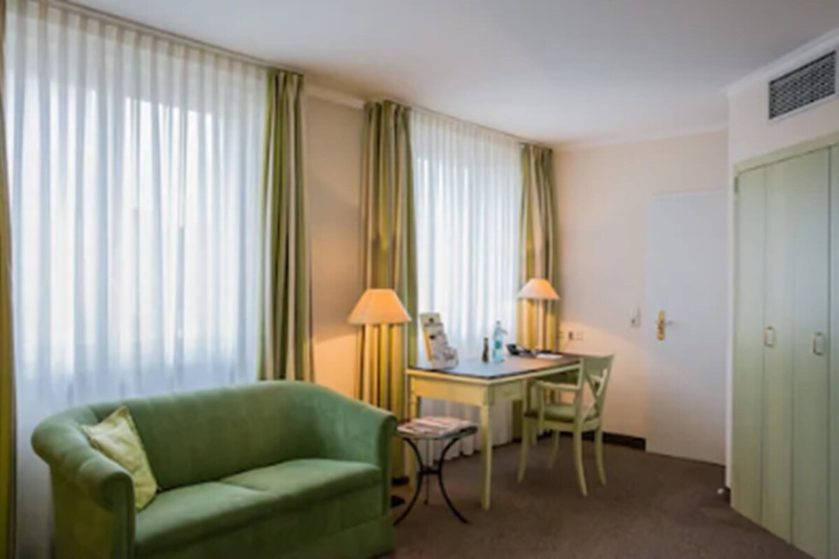 A hotel room with a green sofa, a desk with a lamp, a chair, and a wardrobe.