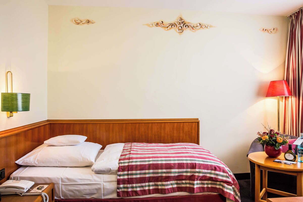 A hotel room with a wooden headboard, a striped bedspread, a bedside table with a lamp, a phone, and a small table with a vase of flowers.