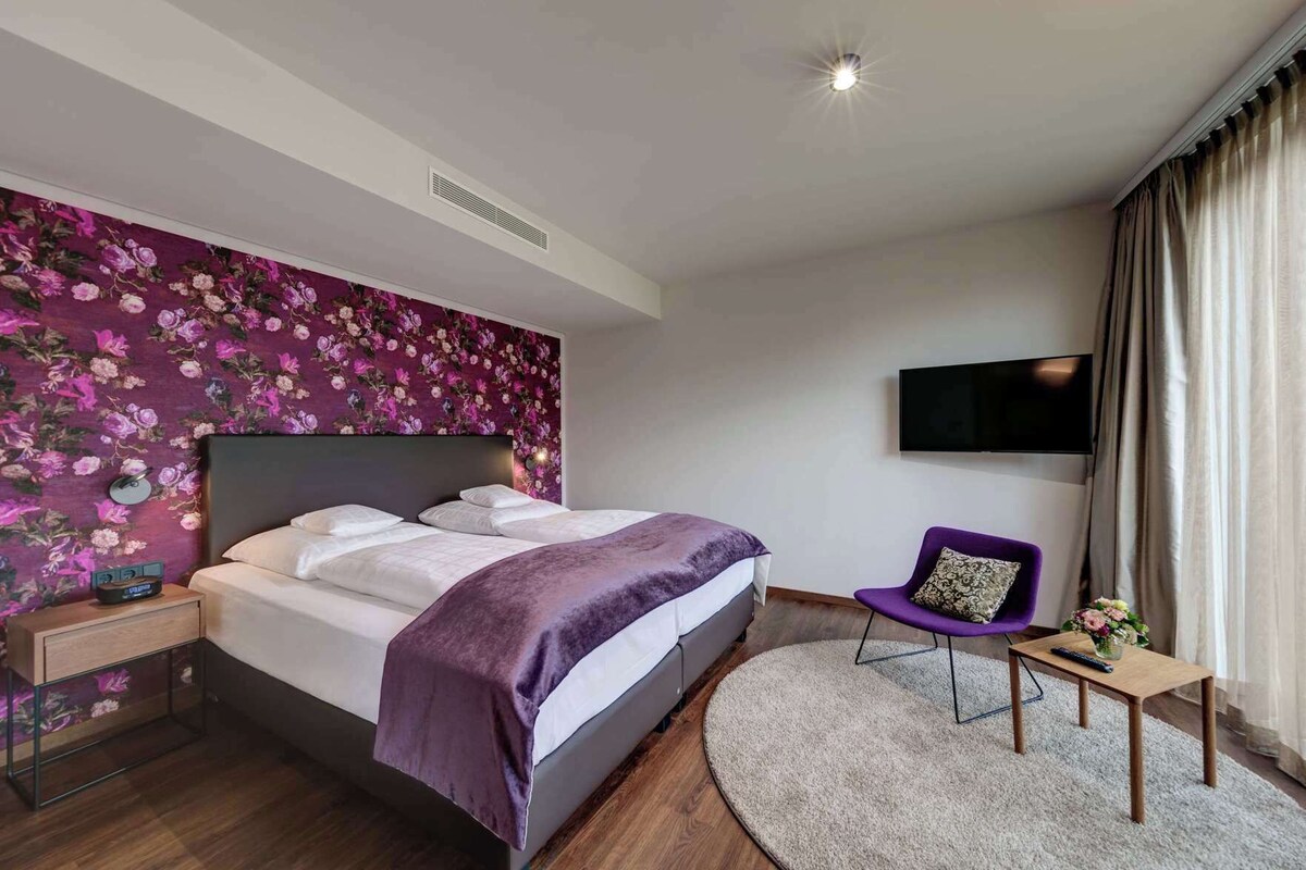 A hotel room with a bed, a TV, a purple chair, a small table, and a floral wall.