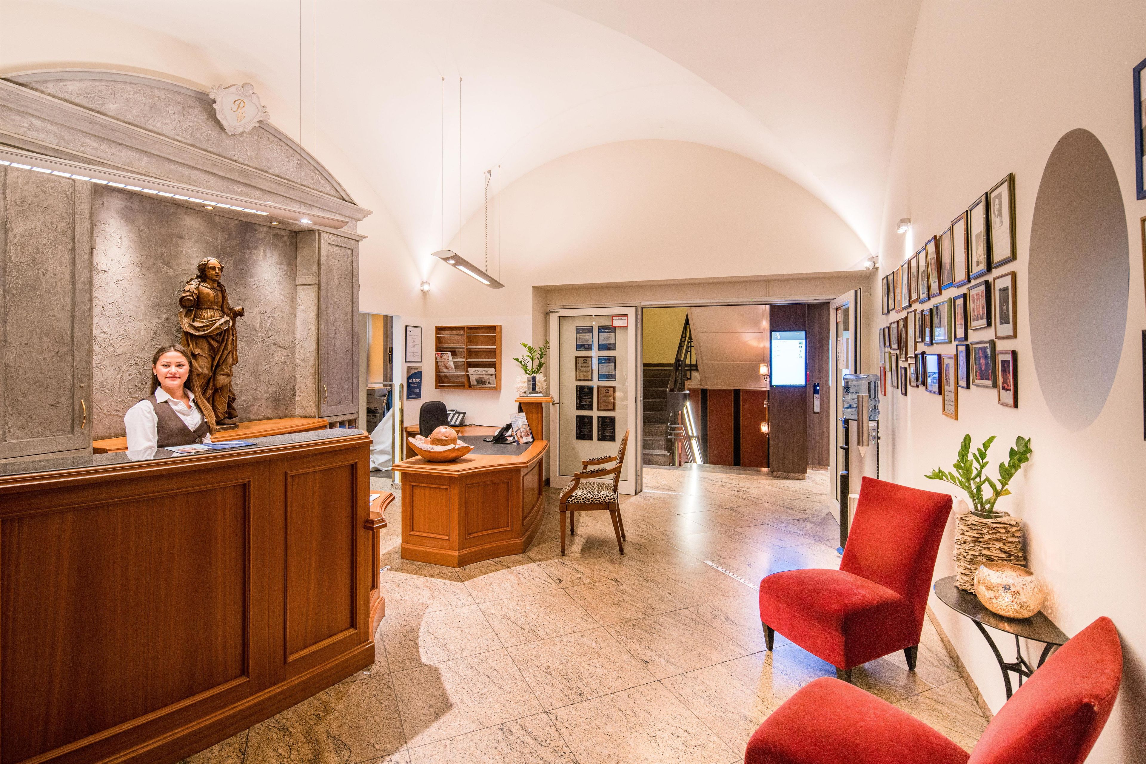 A reception area with a receptionist, a statue, a reception desk, a chair, and a wall with framed pictures.