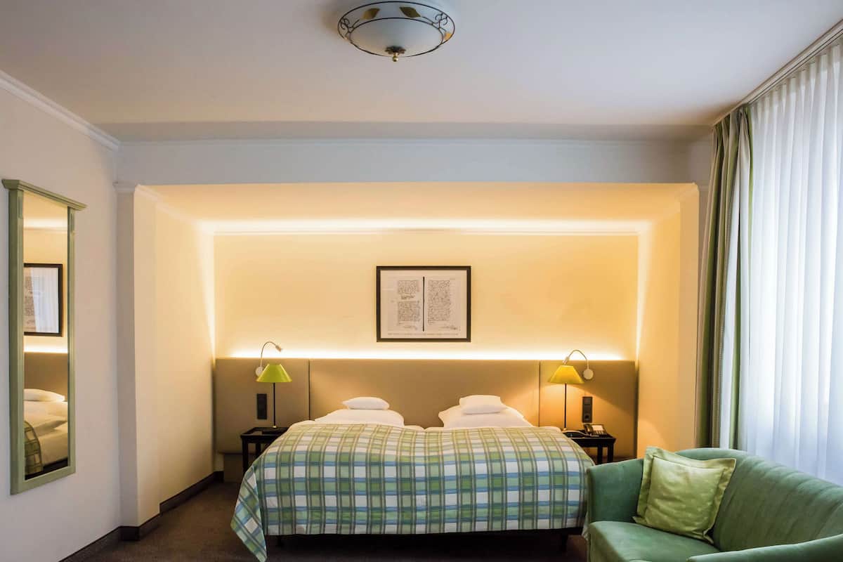 A hotel room with a large bed, two bedside lamps, a green armchair, and a framed picture on the wall.