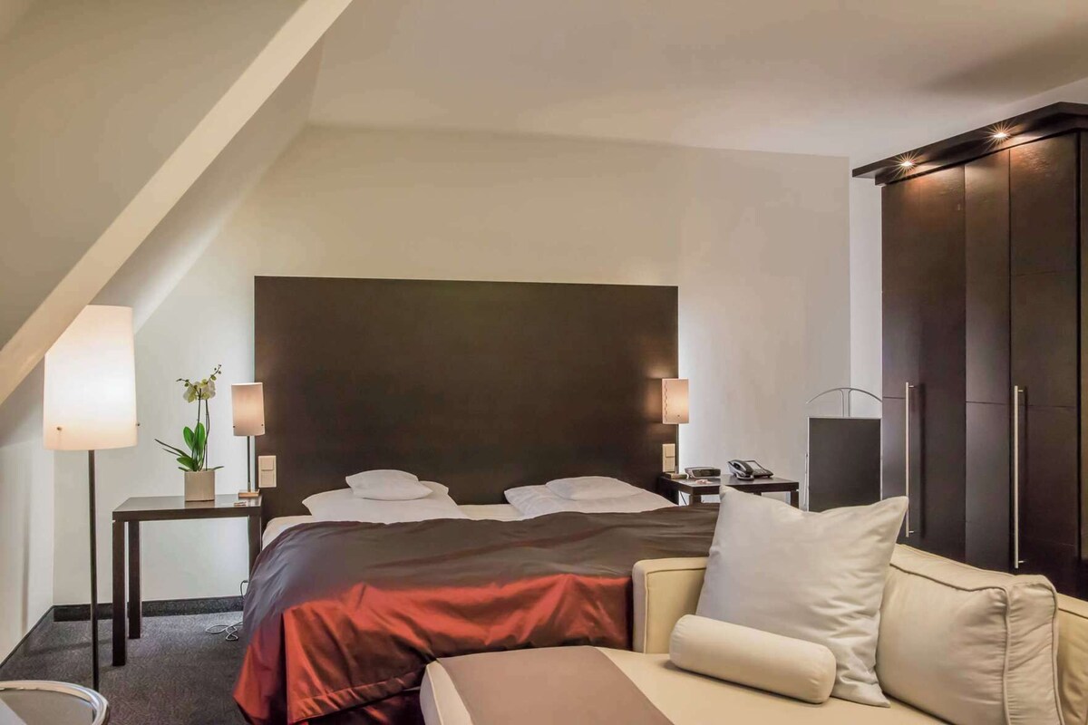 A hotel room with a bed, bedside tables, a sofa, a wardrobe, and a lamp.