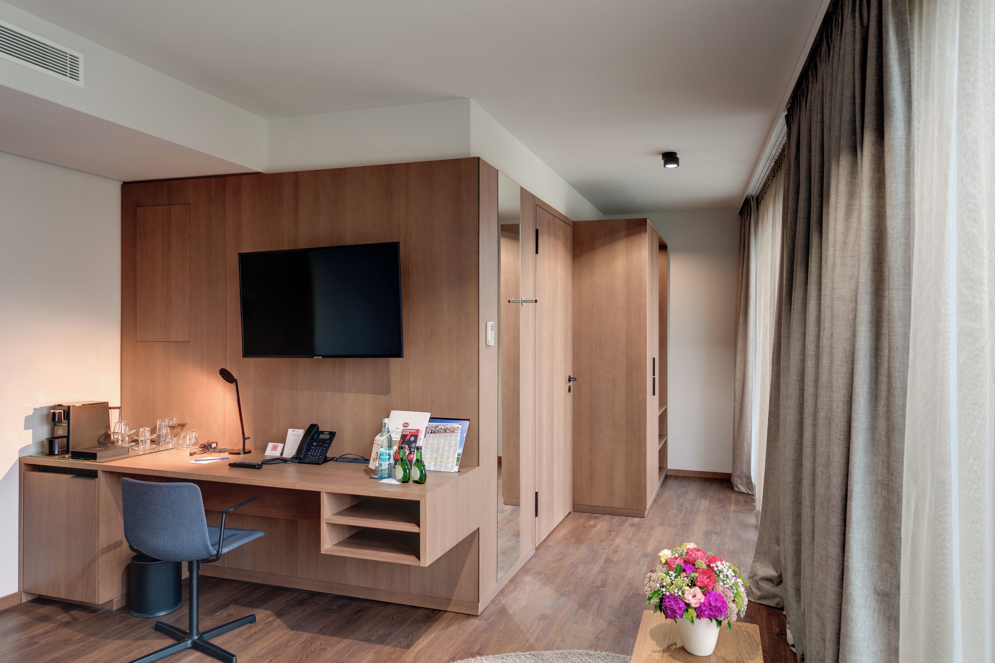 Habitación de hotel moderna con escritorio de madera, televisor de pantalla plana, una silla, máquina de café y una pequeña mesa con un jarrón de flores.