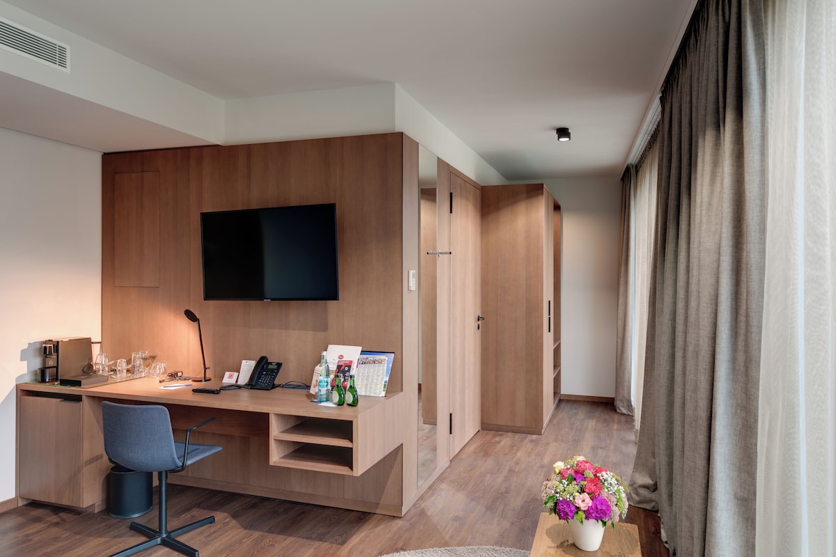 A modern hotel room with a wooden desk, a flat-screen TV, a chair, a coffee machine, and a small table with a vase of flowers.