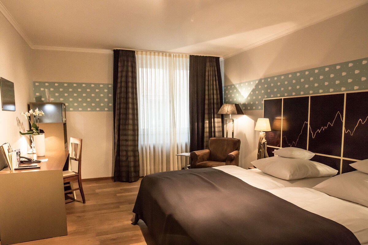 A hotel room with a bed, a desk, a chair, a lamp, and a wall with a pattern.