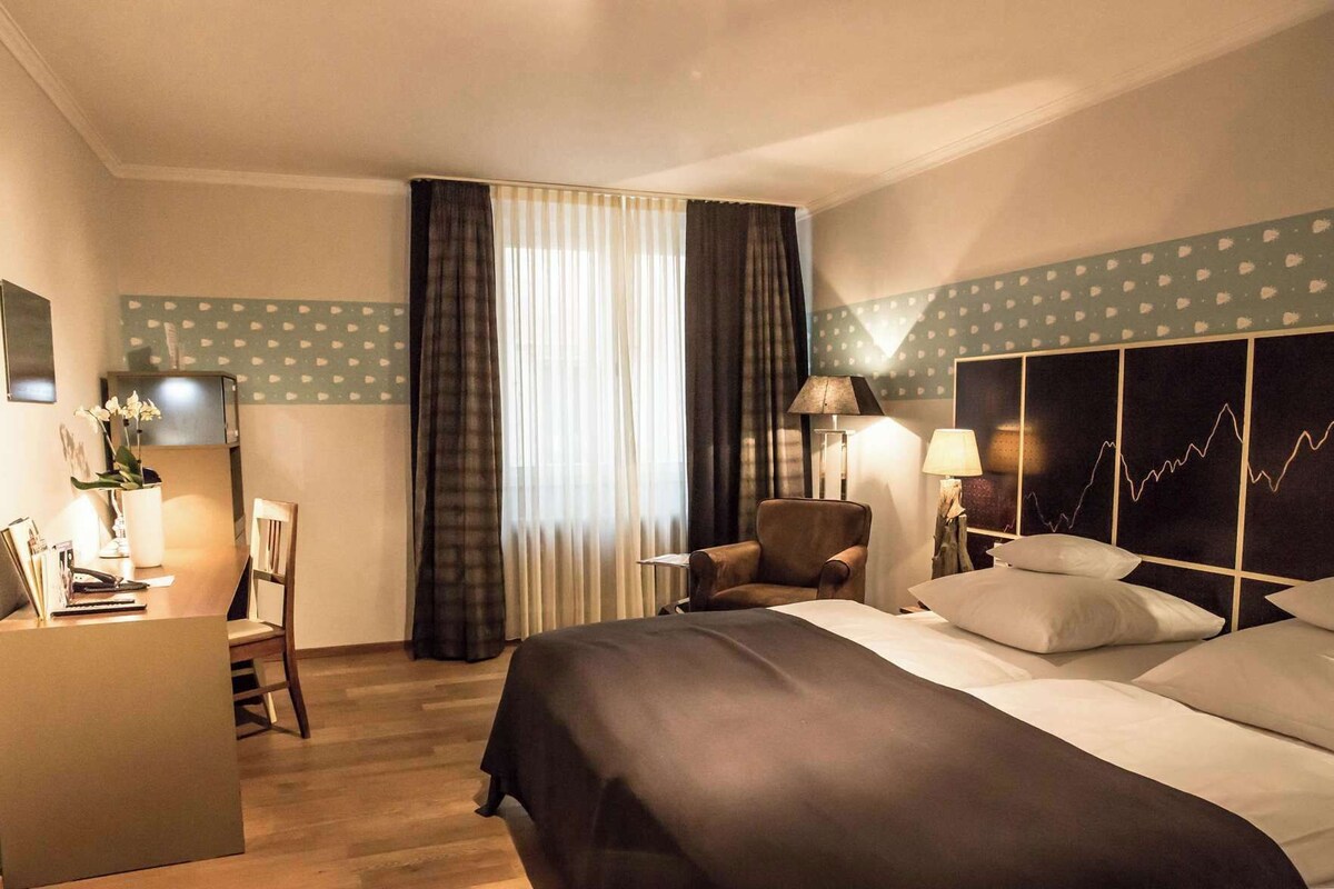 A hotel room with a bed, a desk, a chair, a lamp, and a wall with a pattern.