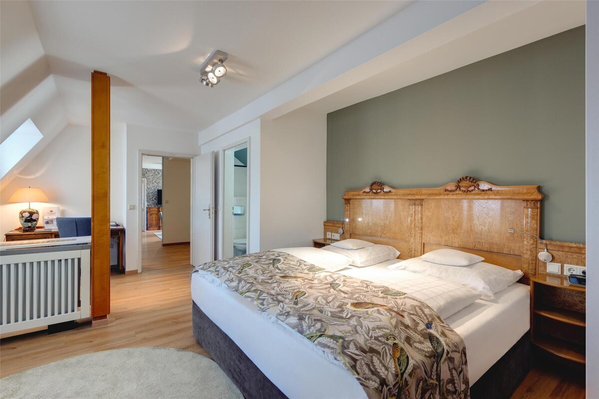 A bedroom with a large bed, a wooden headboard, a radiator, a desk with a lamp, and a view of a bathroom.