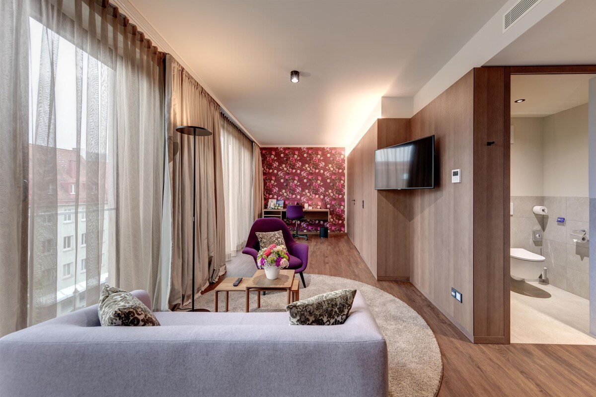 A modern living room with a sofa, a coffee table, and a purple armchair. There is a TV mounted on the wall and a bathroom visible through an open door.