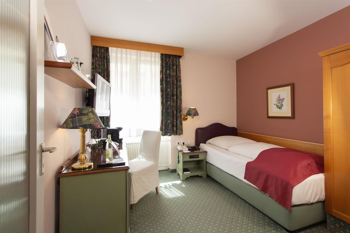 A hotel room with a bed, a desk with a lamp, a chair, a nightstand, and a window with curtains.