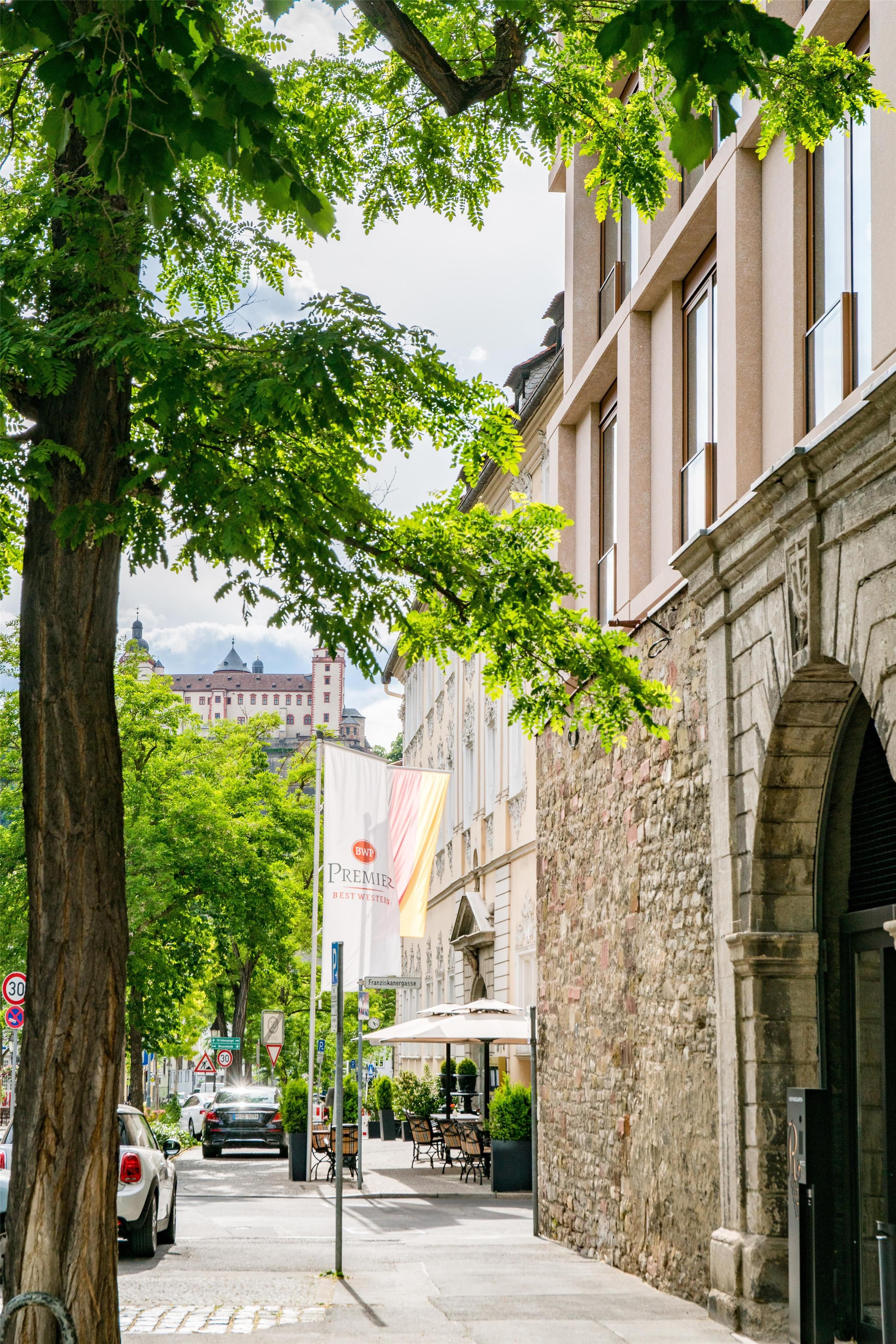 A street with a tree-lined sidewalk, parked cars, and a building with a stone facade and an archway.