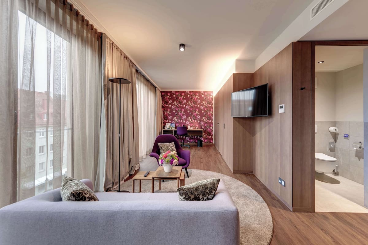 A modern hotel room with a sofa, a small table with a vase, a purple armchair, a TV mounted on the wall, and a bathroom visible through an open door.