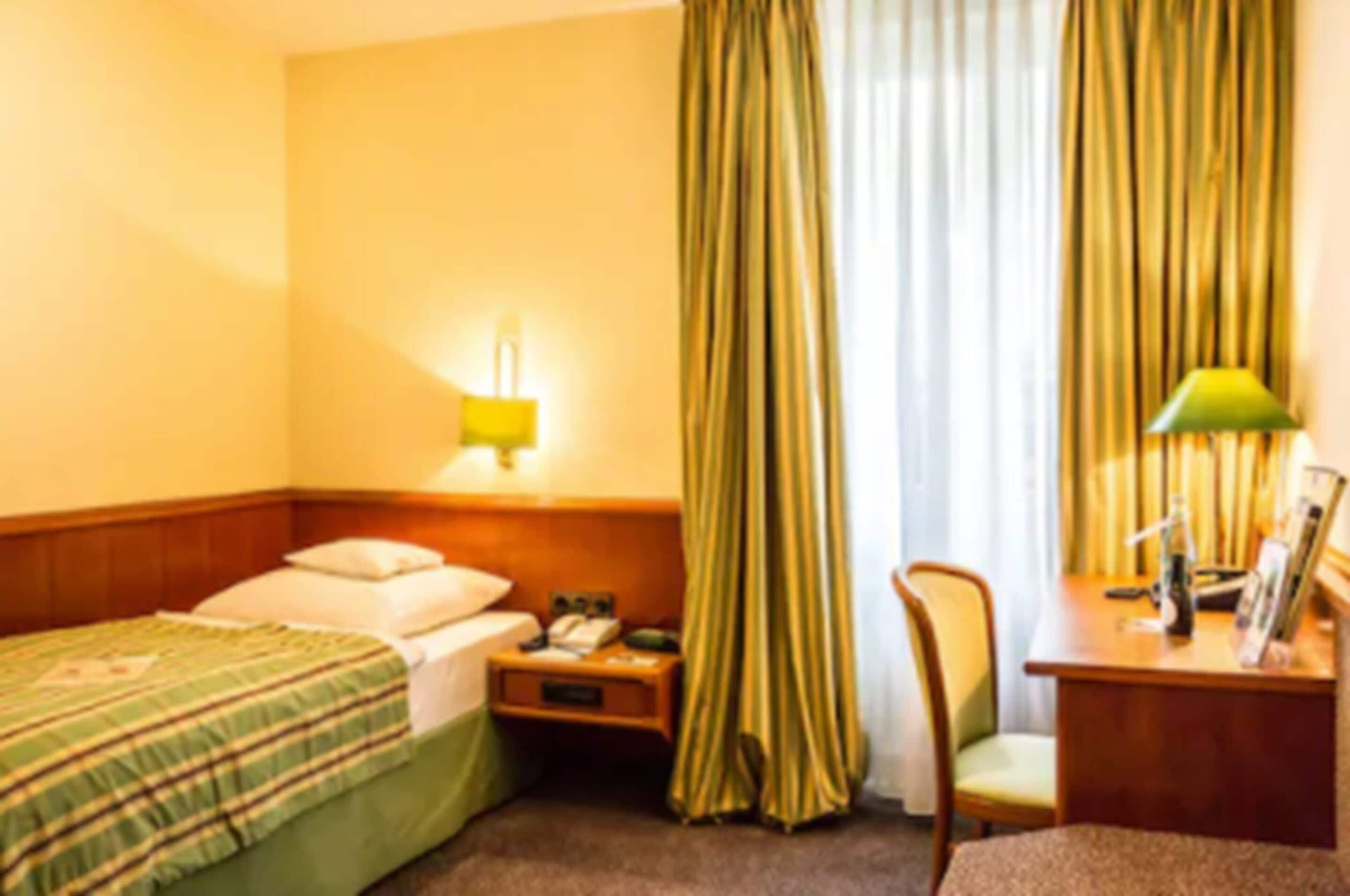 A hotel room with a bed, a nightstand, a desk with a lamp, and a chair.