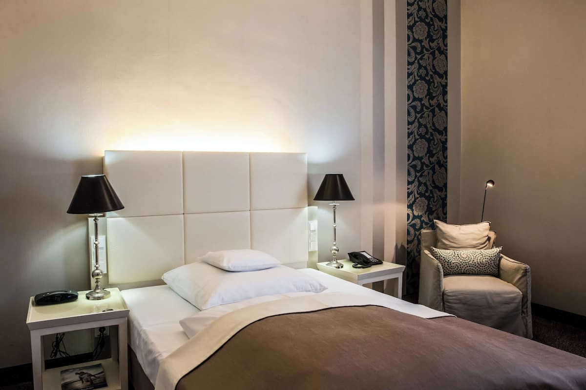 A hotel room with a bed, two bedside tables with lamps, a chair, and a patterned wallpaper.