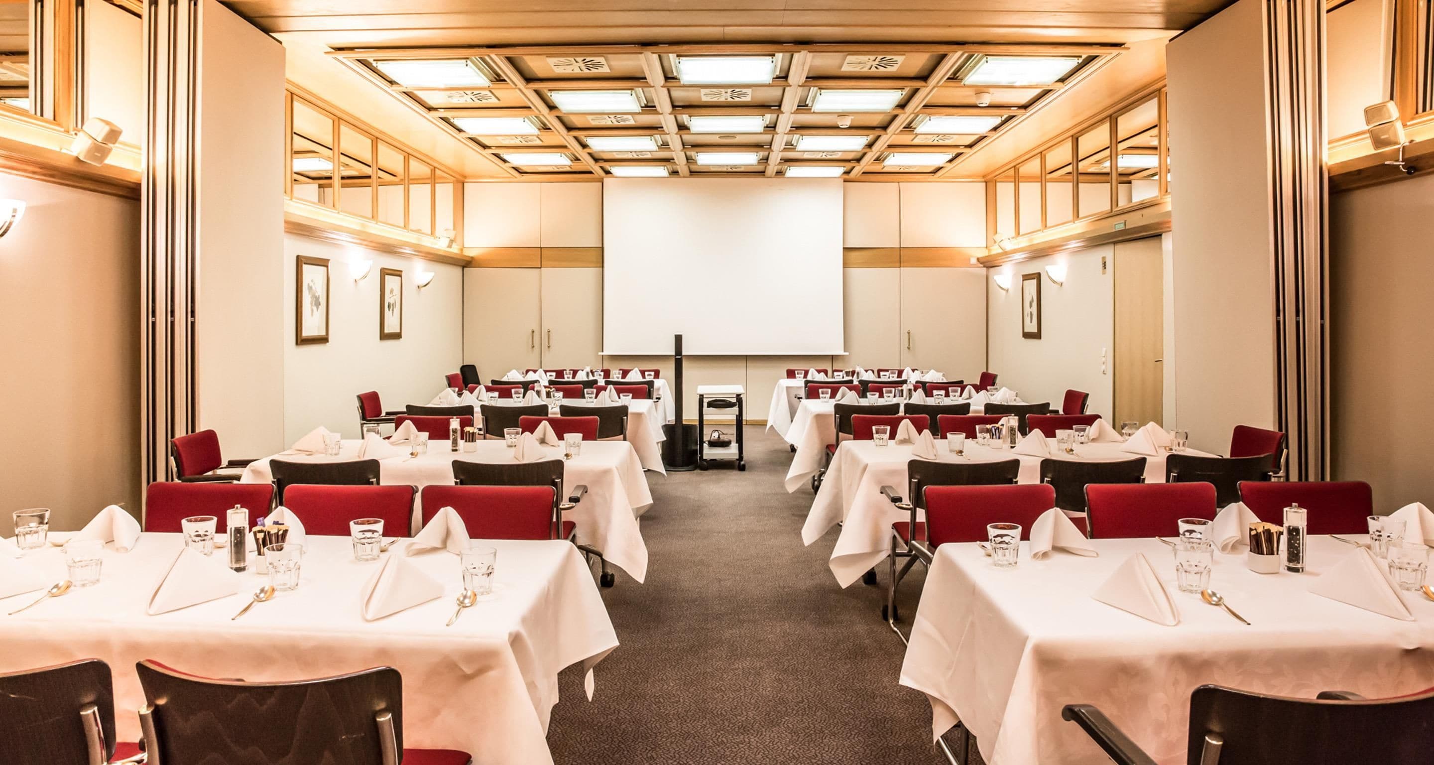 Une salle de conférence avec des tables disposées en rangées, équipée de chaises, de tables et d’un écran de projection.