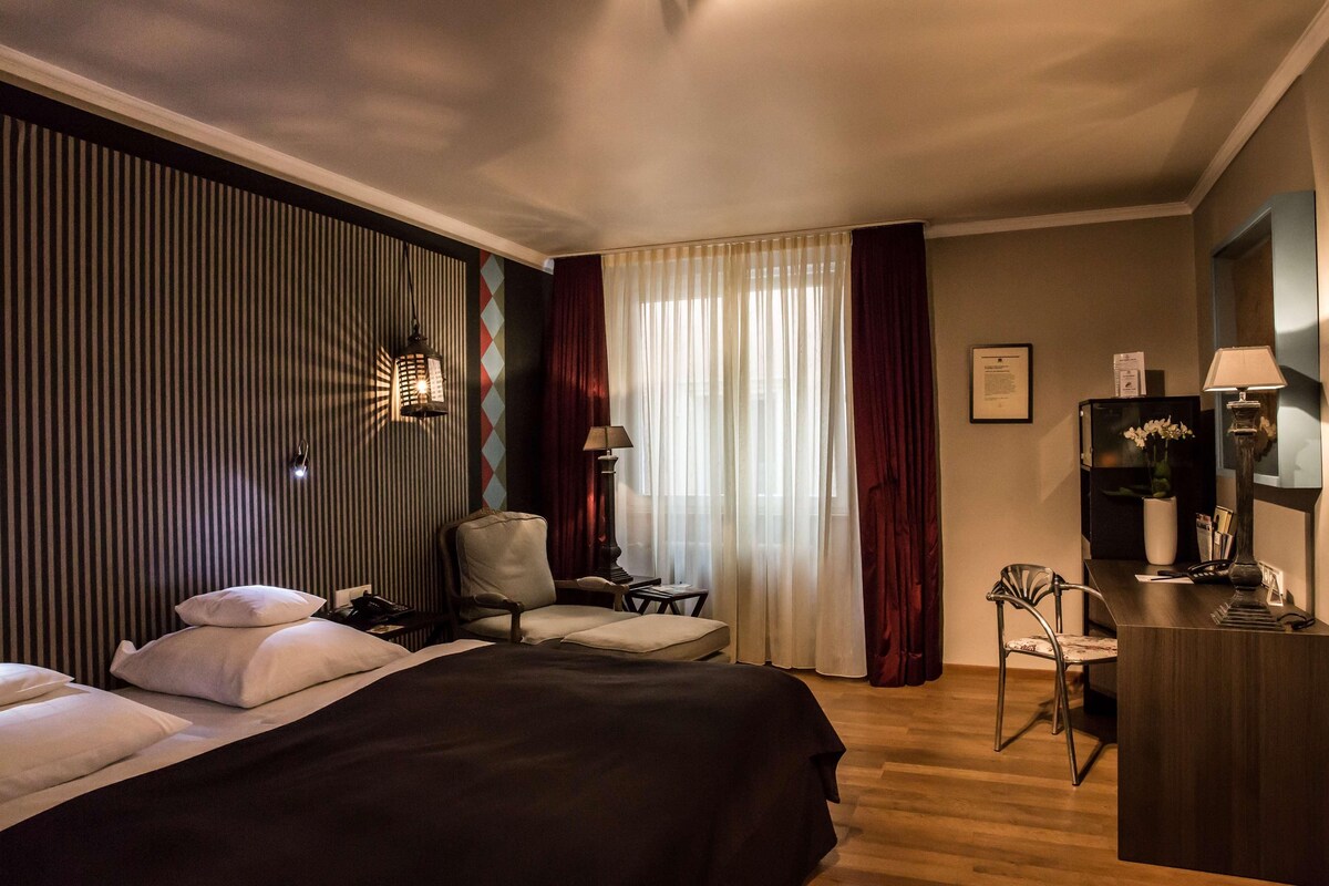 A hotel room with a large bed, a desk, a chair, and a window with curtains.