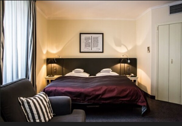 Habitación de hotel con una cama grande, dos mesitas de noche, cabecera, un cuadro enmarcado en la pared, una ventana con cortinas y una puerta.
