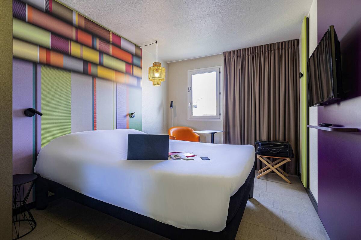 Ein Hotelzimmer mit einem großen Bett, einem Schreibtisch mit Stuhl, einem Fernseher und einem Fenster mit Vorhängen.