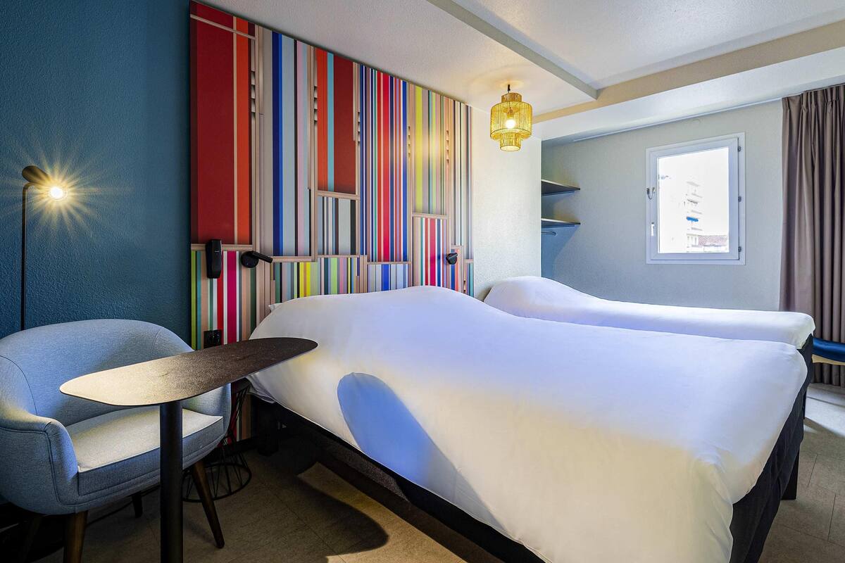 Une chambre d’hôtel avec un lit, une chaise, une petite table, un mur à rayures colorées, une fenêtre et un luminaire suspendu.