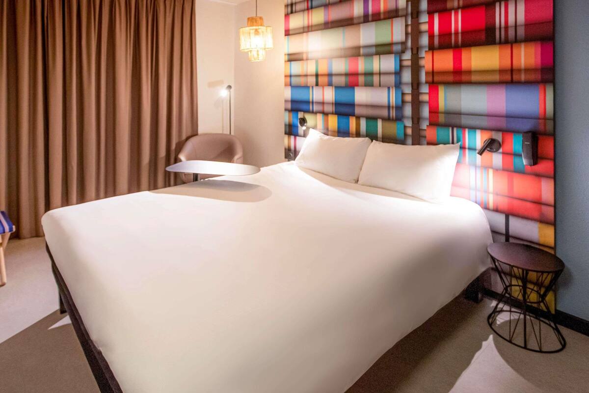 Une chambre d’hôtel avec un grand lit, une chaise, une petite table et un mur aux couleurs vives.