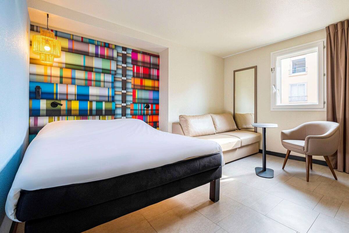 Une chambre d’hôtel comprenant un lit, un canapé, une chaise, une petite table, une décoration murale colorée, une fenêtre avec des rideaux et un miroir.