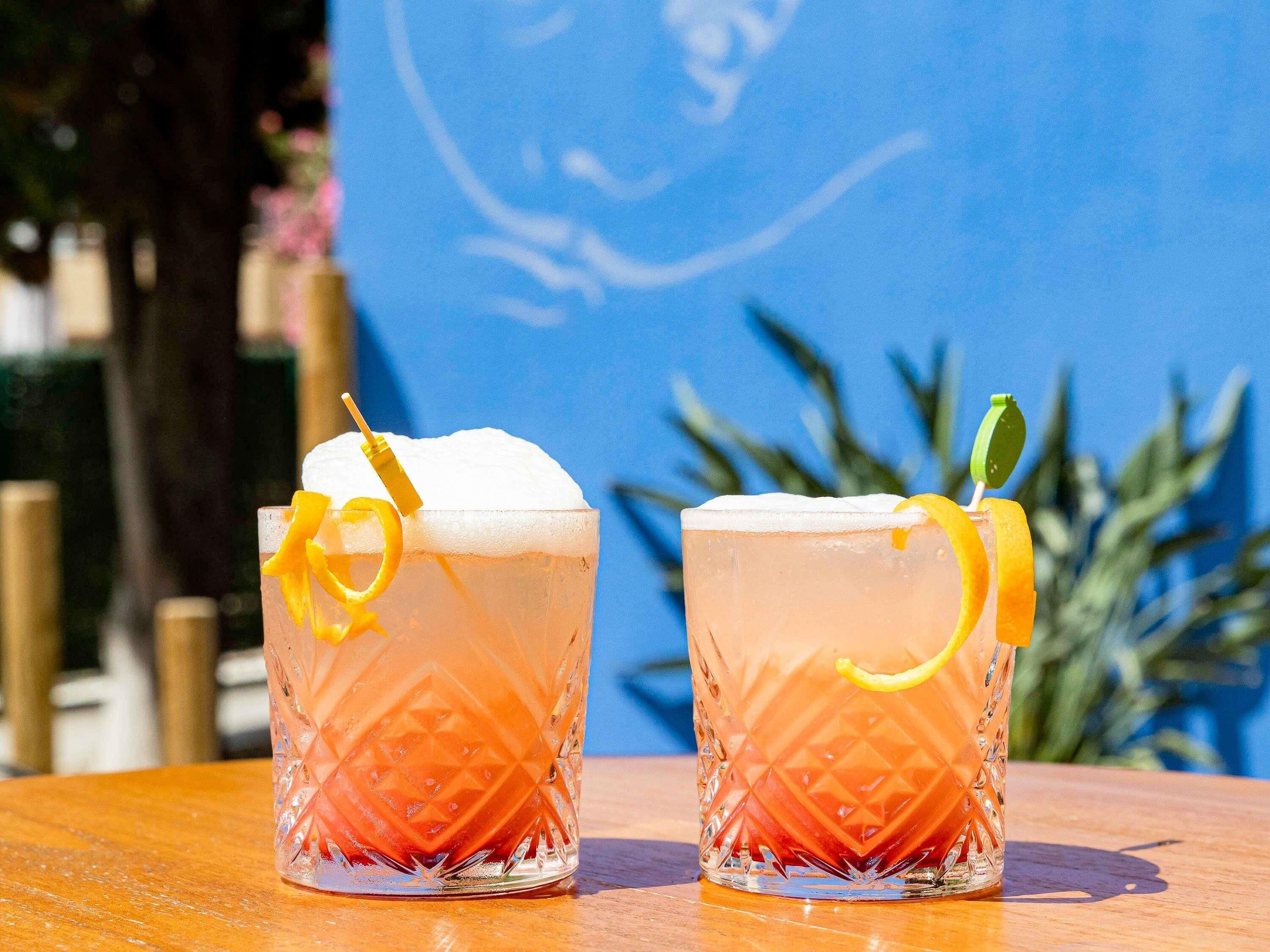 Two glasses of a pink beverage with garnishes on a wooden table against a blue background.