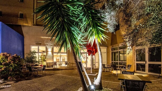 An outdoor seating area with tables and chairs, a palm tree, and a building with large windows in the background. - ibis Styles Perpignan Centre Gare (Perpignan)
