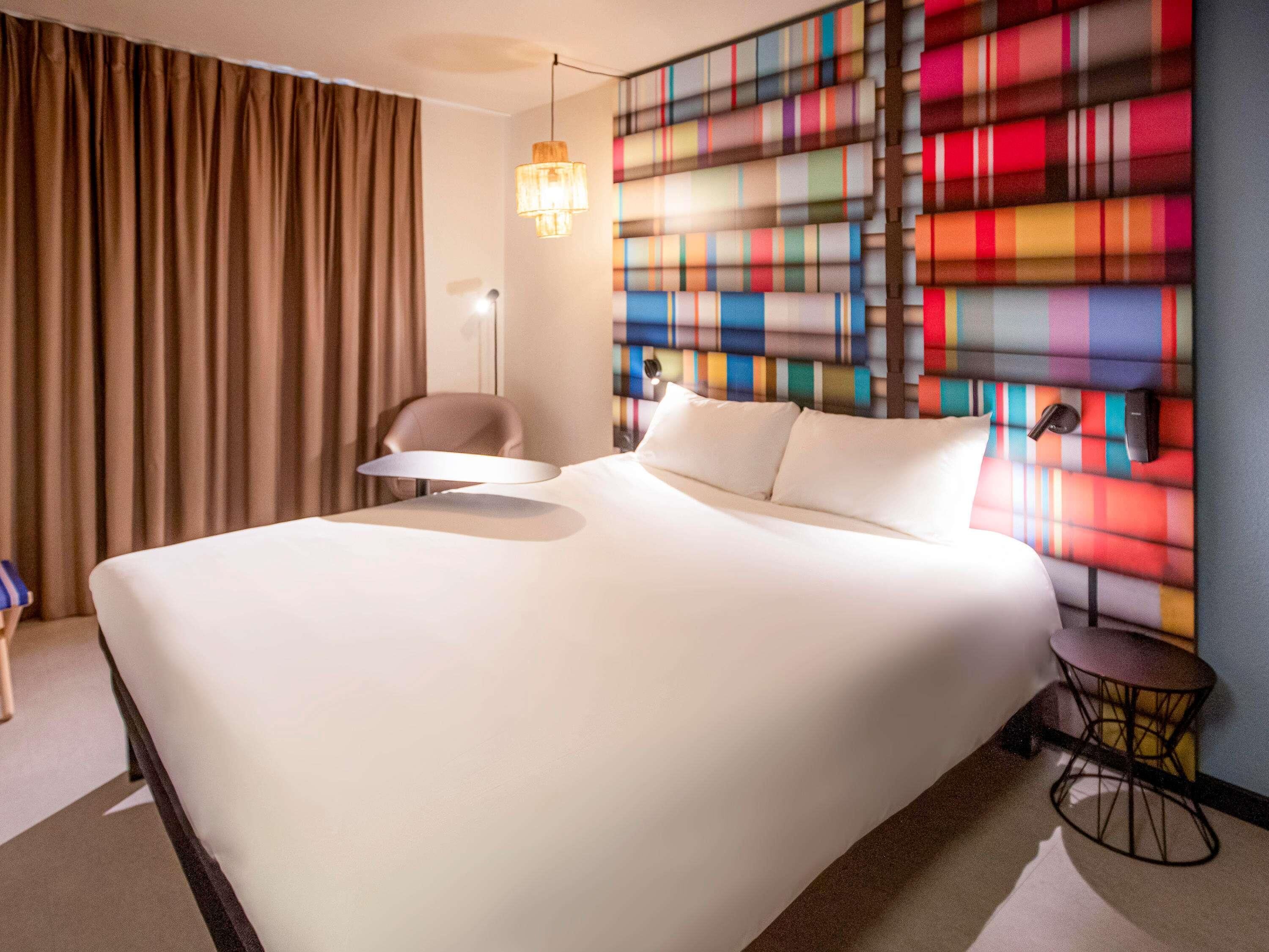 A hotel room with a large bed, a chair, a small table, and a colorful wall.