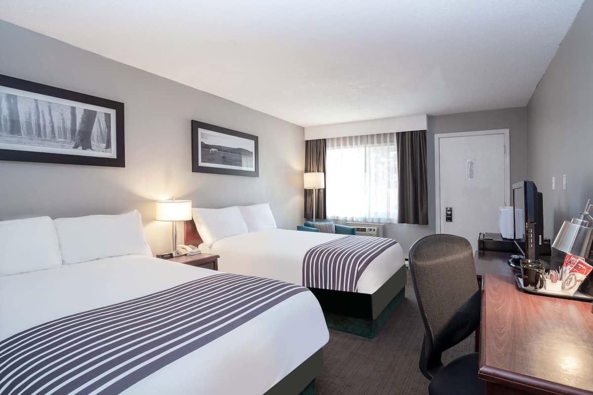 A hotel room with two beds, a desk with a computer, a chair, a coffee machine, and a window with curtains.