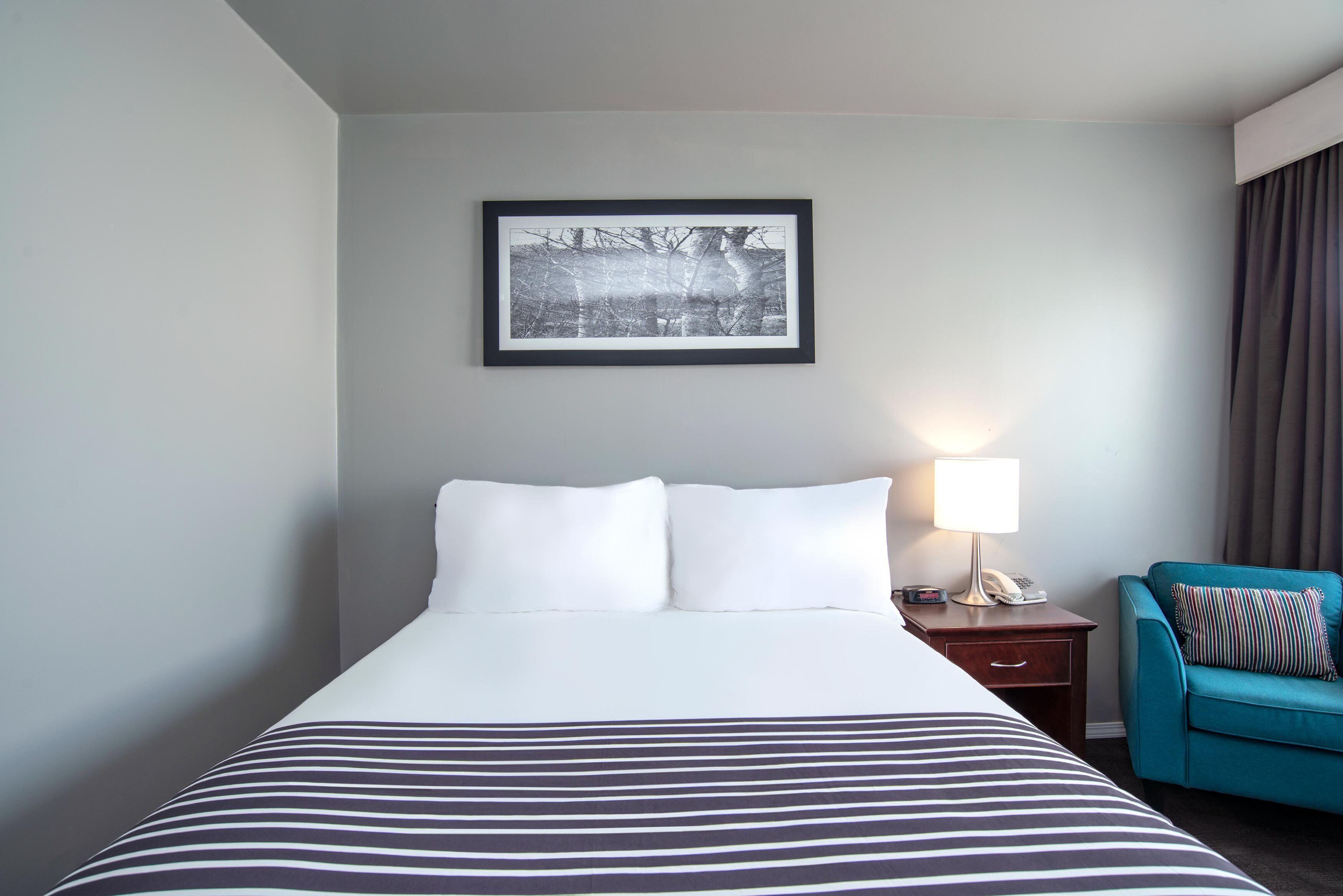 Habitación de hotel con una cama grande, un cuadro en la pared, una lámpara de noche y un sillón azul turquesa.