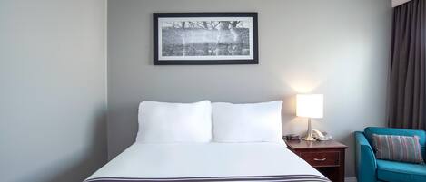 Habitación de hotel con una cama grande, un cuadro en la pared, una lámpara de noche y un sillón azul turquesa.
