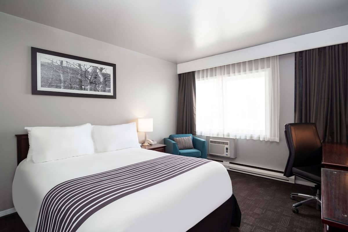 A hotel room with a bed, a desk, a chair, a lamp, an air conditioner, a window with curtains, and a framed picture on the wall.