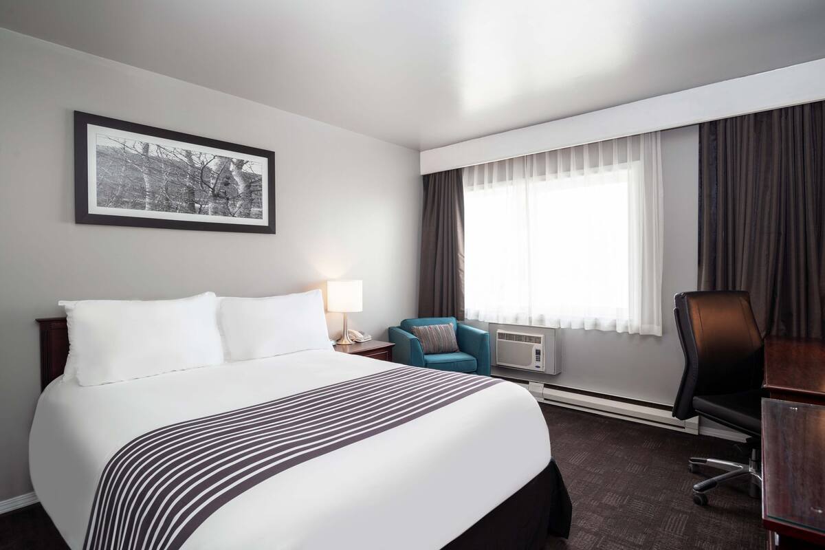 A hotel room with a large bed, a desk, a chair, an air conditioning unit, and a window with curtains.