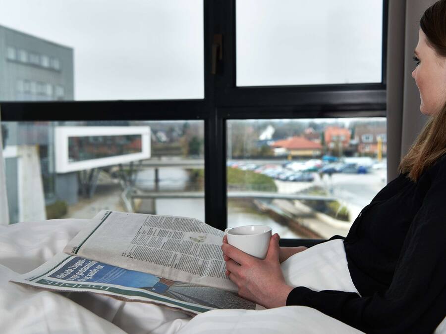 Una persona leyendo un periódico en una cama con vista a un estacionamiento y edificios a través de la ventana.