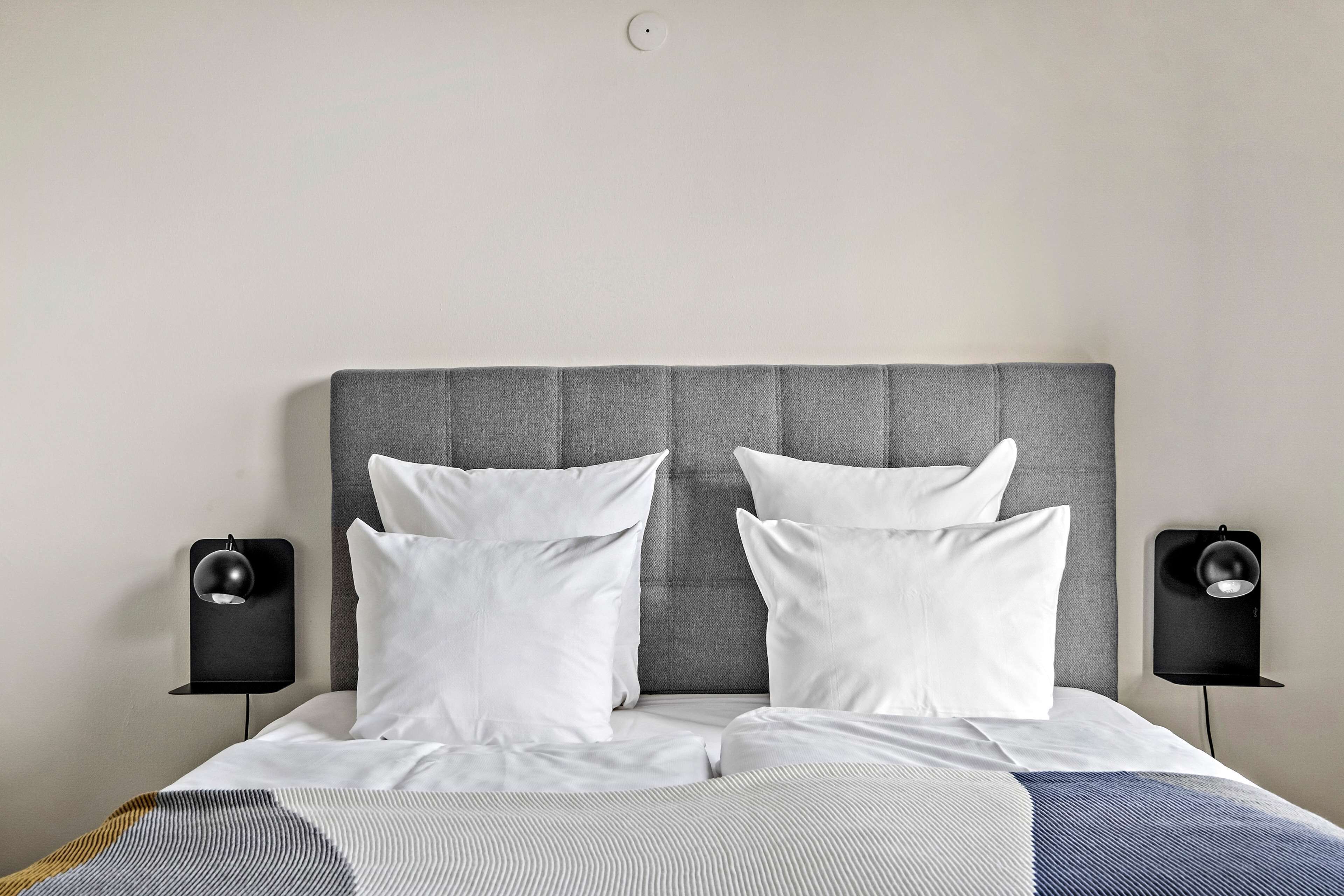 Een net opgemaakt bed met witte lakens en een grijze hoofdbord, aan weerszijden verlicht door twee zwarte bedlampen.