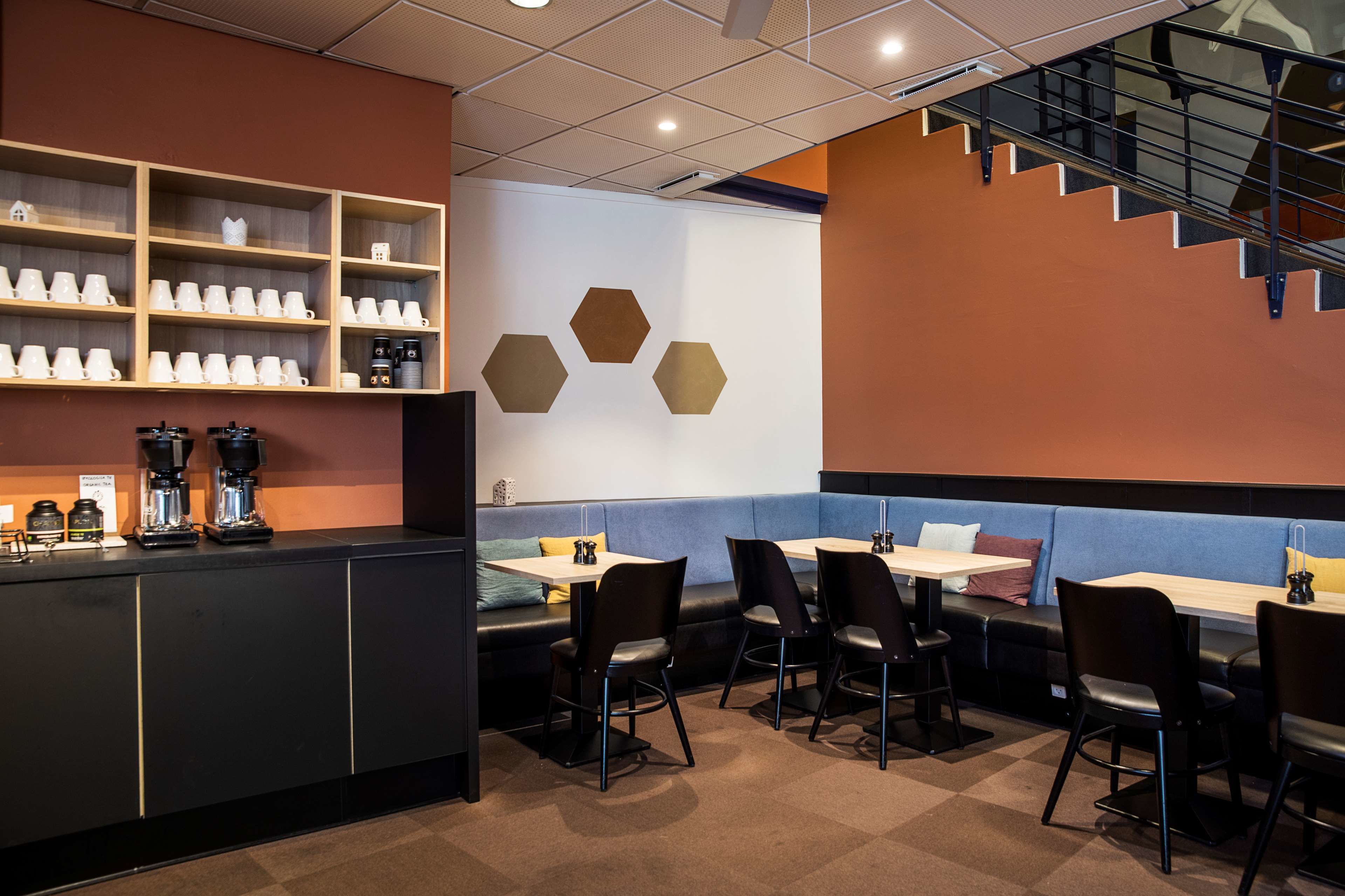 Un café moderno con un mostrador que exhibe cafeteras y tazas, zona de estar con mesas y sillas, y una escalera.